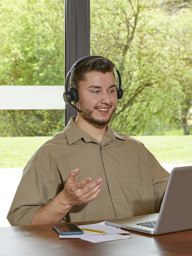 Person sitzt am Tisch und unterhält sich glücklich mit Hilfe des Wireless Office Headsets über ein Laptop.