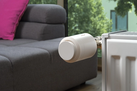In the foreground you can see a smart Hama heating thermostat attached to a radiator. A living room with a dark leather sofa can be seen in the background.